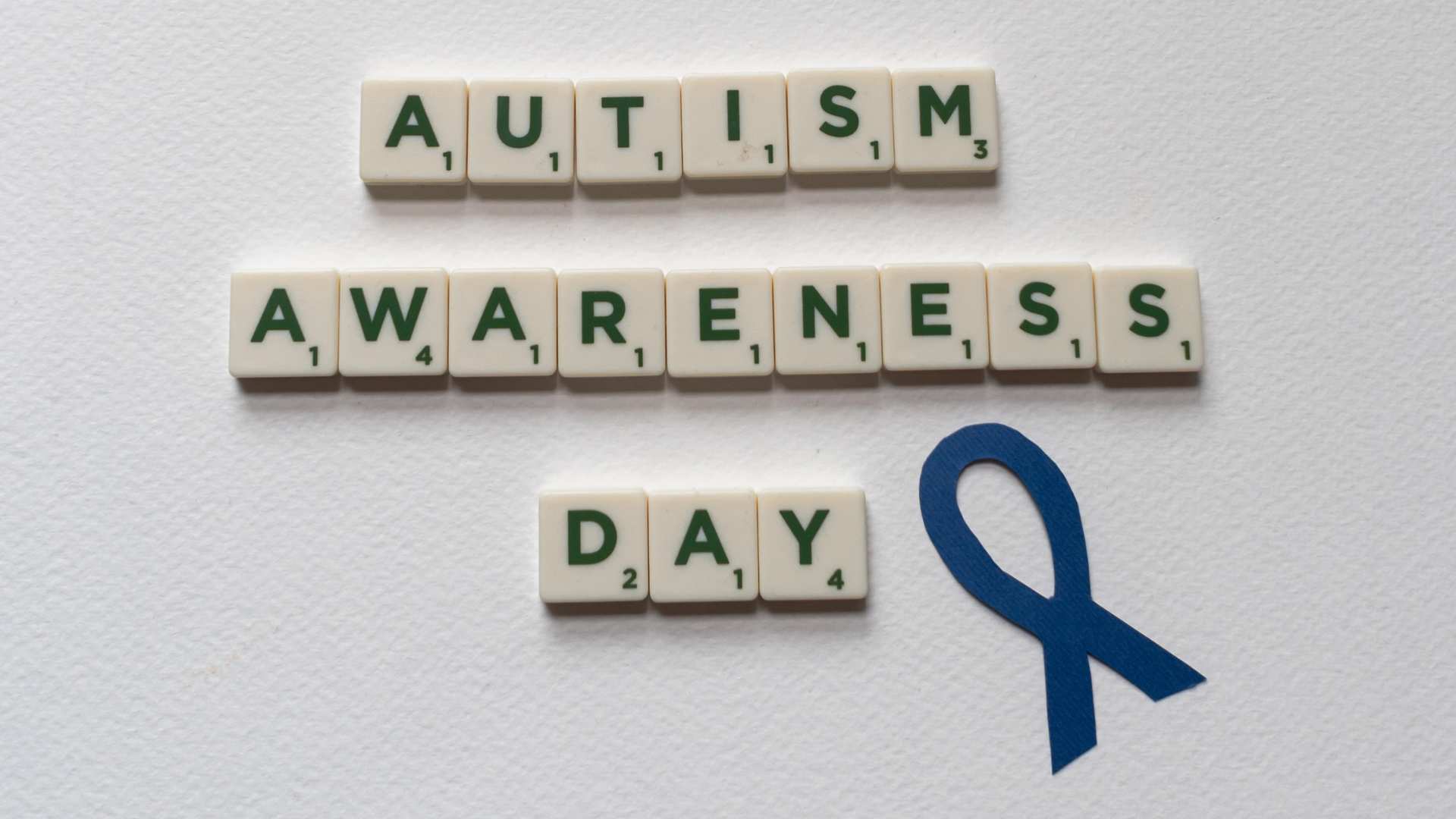 The Meaning Behind World Autism Awareness Day A blue rainbow with clouds above letter tiles spelling Autism Awareness Day and a blue ribbon