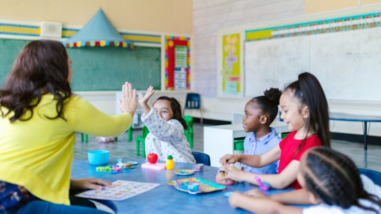 ABA professional high-fives a young girl during an in-school ABA therapy session while other children engage with learning materials at a classroom table